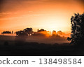 A peaceful summer morning landscape featuring a glowing sunrise over a mist-covered rural field.  138498584