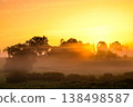 A peaceful summer morning landscape featuring a glowing sunrise over a mist-covered rural field.  138498587