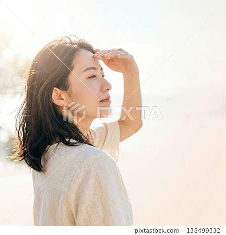 A woman shielding her eyes from the sun with her hands, an image representing protection against strong sunlight and summer UV rays. 138499332