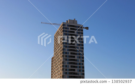 The beam of a high-rise construction crane is visible from behind a multi-story building. Modern geometric architecture The beam of a high-rise construction crane is visible from behind a multi-story building. Modern geometric architecture 138502937