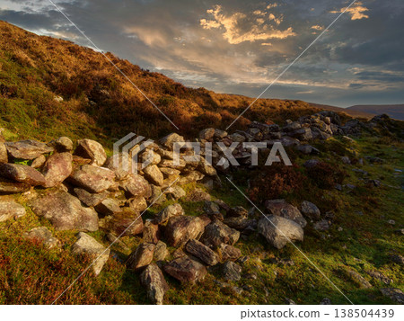 An old stone wall sits on a grassy hillside under a cloudy sky during sunset. An old stone wall sits on a grassy hillside under a cloudy sky during sunset. 138504439