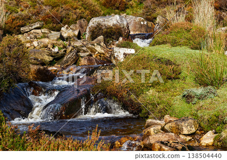 Small stream flowing over rocks and boulders through grassy terrain with low shrubs and patches of vegetation. Small stream flowing over rocks and boulders through grassy terrain with low shrubs and patches of vegetation. 138504440