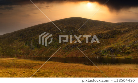 A vast mountain looms against the backdrop of a vibrant orange sunset sky. In the foreground, a calm, dark lake rests at its base, surrounded by golden and green moorland vegetation. A vast mountain looms against the backdrop of a vibrant orange sunset sky. In the foreground, a calm, dark lake rests at its base, surrounded by golden and green moorland vegetation. 138504450