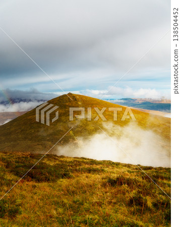 Golden sun shines on a vast mountain, its expansive slopes covered in rich grass and rising mist. The cloudy sky shows glimpses of blue, suggesting changing weather over this grand natural landscape. Golden sun shines on a vast mountain, its expansive slopes covered in rich grass and rising mist. The cloudy sky shows glimpses of blue, suggesting changing weather over this grand natural landscape. 138504452