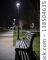 Empty park bench resting beside a paved pathway with modern streetlights creating a peaceful ambiance, offering a quiet space for contemplation or relaxation outdoors 138504635