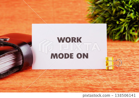 White card declaring work mode on rests firmly on a textured orange desktop. A closed black notebook and a lively green plant complete the productive scene White card declaring work mode on rests firmly on a textured orange desktop. A closed black notebook and a lively green plant complete the productive scene 138506141