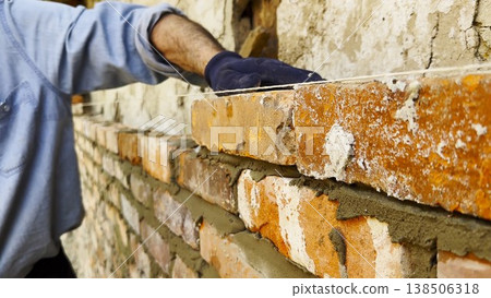 Adult male hands in protective gloves apply cement with a trowel and place bricks to build brick wall for house. The worker carefully spreads mortar and sets each brick during the construction process Adult male hands in protective gloves apply cement with a trowel and place bricks to build brick wall for house. The worker carefully spreads mortar and sets each brick during the construction process 138506318