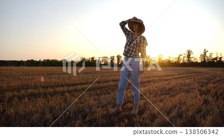 Adult agronomist in straw hat exploring barley on meadow at sunset. Female farmer examining ripe wheat at field on dusk. Beautiful scenic landscape. Concept of agriculture and agronomy business Adult agronomist in straw hat exploring barley on meadow at sunset. Female farmer examining ripe wheat at field on dusk. Beautiful scenic landscape. Concept of agriculture and agronomy business 138506342