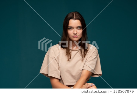 Young woman with brown hair and light eyes looking at camera against teal background, presenting neutral expression Young woman with brown hair and light eyes looking at camera against teal background, presenting neutral expression 138507079