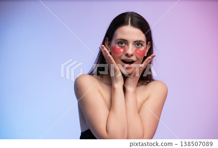 Young woman applying red hydrogel eye patches for skincare treatment on her face, experiencing self-care and beauty with a surprised expression and glowing skin 138507089