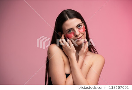 Young woman applying under eye hydrogel patches for hydration and skincare, practicing self-care and beauty routine with pink background 138507091