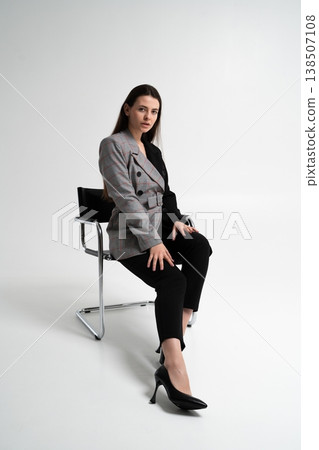 Woman wearing a stylish two-tone blazer and black trousers, sitting confidently on a modern chair in a studio, representing business, fashion, and corporate success Woman wearing a stylish two-tone blazer and black trousers, sitting confidently on a modern chair in a studio, representing business, fashion, and corporate success 138507108