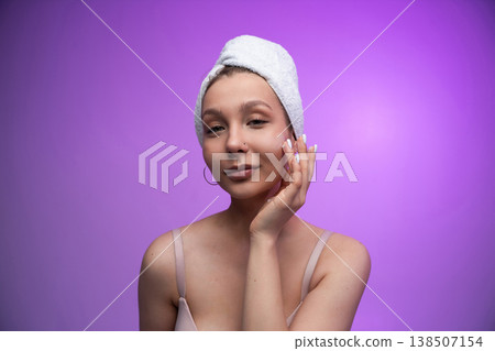 Young woman applying face cream, pampering facial skin, moisturizing after showering for wellness and beauty, embracing self care against a purple background 138507154