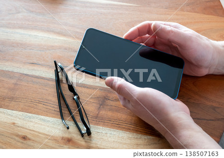 Man take new mobile phone in the hands on the wood table. Smartphone and glasses for work in office. Man take new mobile phone in the hands on the wood table. Smartphone and glasses for work in office. 138507163