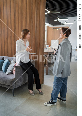 Female professionals facing each other having conversation in bright modern office interior. Conversation, standing, teamwork, office, businesswomen. Female professionals facing each other having conversation in bright modern office interior. Conversation, standing, teamwork, office, businesswomen. 138508384