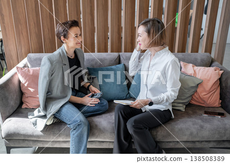 Two women in business attire talking and looking at laptop on sofa in modern office. Professional discussion, business, coworking, teamwork, office. 138508389