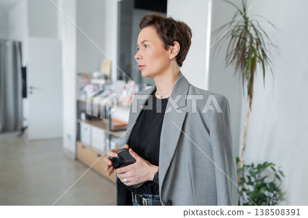 Businesswoman smiling and holding folder while talking to colleague in bright office with green plant. Smiling, talking, businesswoman, office, folder, friendly. 138508391