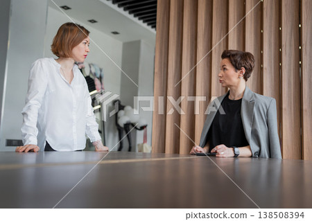 Businesswomen discussing and smiling at worktable in well-lit modern office, both standing. Discussion, smiling, teamwork, business, office. Businesswomen discussing and smiling at worktable in well-lit modern office, both standing. Discussion, smiling, teamwork, business, office. 138508394
