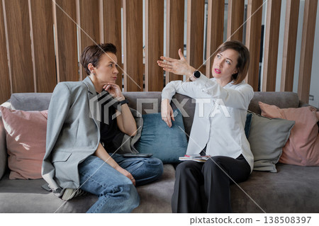 Two women on sofa discussing and laughing while talking animatedly in creative office. Teamwork, laughter, discussion, office, business. Two women on sofa discussing and laughing while talking animatedly in creative office. Teamwork, laughter, discussion, office, business. 138508397