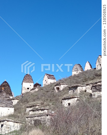 View of the City of the Dead, Dargavs necropolis, North Ossetia, Russia. Ancient stone crypts scattered across a green mountain slope under a blue sky View of the City of the Dead, Dargavs necropolis, North Ossetia, Russia. Ancient stone crypts scattered across a green mountain slope under a blue sky 138508925