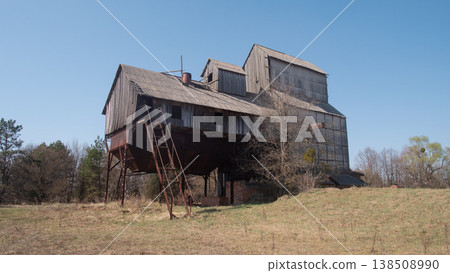 Abandoned Wooden Farmhouse In Field With Rusted External Stairs, Boarded Windows, Overgrown Grass, Surrounding Pine Trees Under Clear Blue Sky, Atmosphere Of Isolation And Quiet, Visible 138508990