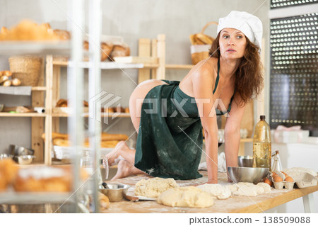 Sexy female baker in apron on naked body posing sexy on knees on kitchen table of bakery Sexy female baker in apron on naked body posing sexy on knees on kitchen table of bakery 138509088