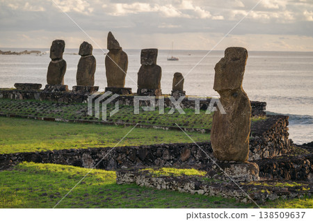 Majestic moai statue stands stoic on grassy shore of rapa nui easter island looking over peaceful ocean water beside an ancient stone monument under grey cloudy sky 138509637