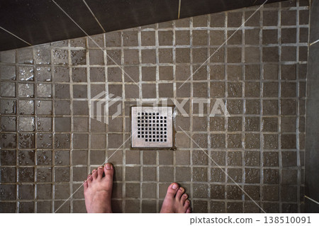 Looking down to hotel room shower drain grid, wet tiles on floor, man feet visible in lower part 138510091
