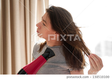 A woman blow-drying her hair, soft morning sunlight shining through the curtains, a concentrated expression on her face, hands running through chestnut curls, a home routine, relaxed self-care. A woman blow-drying her hair, soft morning sunlight shining through the curtains, a concentrated expression on her face, hands running through chestnut curls, a home routine, relaxed self-care. 138510100