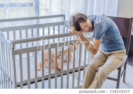 Tired father sitting by baby crib at home watching sleeping infant. Exhausted dad caring for newborn at night. Parenting fatigue, childcare responsibility, love and real family life concept Tired father sitting by baby crib at home watching sleeping infant. Exhausted dad caring for newborn at night. Parenting fatigue, childcare responsibility, love and real family life concept 138510257