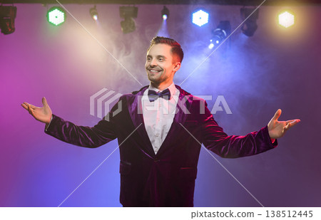 Happy presenter in bow tie on stage under vibrant lights with open arms wide Happy presenter in bow tie on stage under vibrant lights with open arms wide 138512445
