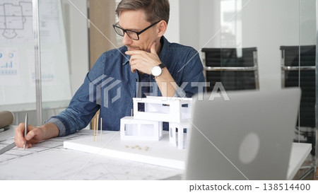 Male architect thinking intently while reviewing architectural blueprints and a 3d scale model of a contemporary house, brainstorming design ideas in a professional office environment Male architect thinking intently while reviewing architectural blueprints and a 3d scale model of a contemporary house, brainstorming design ideas in a professional office environment 138514400
