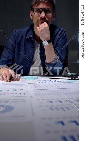 Caucasian man sitting at an office desk in the evening, concentrating and writing notes while surrounded by business documents, charts, graphs, and a laptop Caucasian man sitting at an office desk in the evening, concentrating and writing notes while surrounded by business documents, charts, graphs, and a laptop 138514411