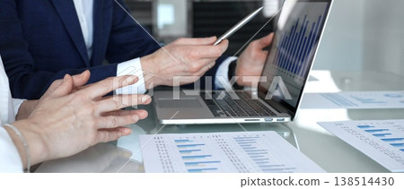 Business professionals collaborating in a modern office, reviewing financial reports, charts, and a laptop on a glass table while discussing data analytics, business strategy, and planning Business professionals collaborating in a modern office, reviewing financial reports, charts, and a laptop on a glass table while discussing data analytics, business strategy, and planning 138514430
