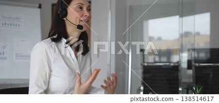 Businesswoman in a white shirt and headset leading a video call from a modern office, presenting with a tablet and gesturing as she discusses strategy and trains remote colleagues 138514617