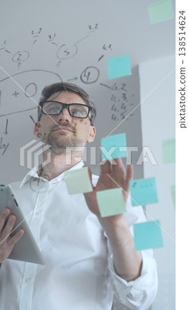 Mature businessman in glasses actively analyzing data and developing new business strategies. Using a digital tablet during a dynamic brainstorming and planning session behind a glass wall with notes Mature businessman in glasses actively analyzing data and developing new business strategies. Using a digital tablet during a dynamic brainstorming and planning session behind a glass wall with notes 138514624