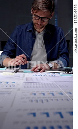 Professional man in glasses working late at night, analyzing financial data on a digital tablet, managing business reports, and extending productivity in a dark office environment Professional man in glasses working late at night, analyzing financial data on a digital tablet, managing business reports, and extending productivity in a dark office environment 138514633