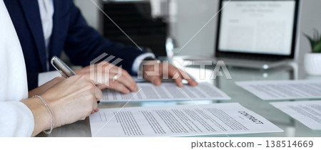 Business people are discussing a contract, reviewing documents and pointing with a pen, sitting at an office table with a laptop, focusing on agreement details and legal paperwork 138514669