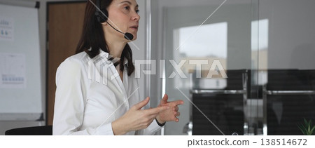 Businesswoman in a white shirt and headset leading a video call from a modern office, presenting with a tablet and gesturing as she discusses strategy and trains remote colleagues Businesswoman in a white shirt and headset leading a video call from a modern office, presenting with a tablet and gesturing as she discusses strategy and trains remote colleagues 138514672