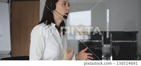 Businesswoman in a white shirt and headset leading a video call from a modern office, presenting with a tablet and gesturing as she discusses strategy and trains remote colleagues 138514691