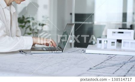 Woman architect working at an office desk, analyzing blueprints, using a laptop, and preparing a modern house model, representing innovative construction and design processes Woman architect working at an office desk, analyzing blueprints, using a laptop, and preparing a modern house model, representing innovative construction and design processes 138514717