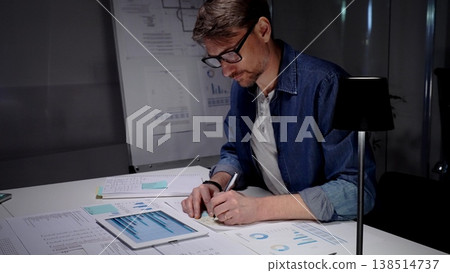 Professional man in glasses working late at night, analyzing financial data on a digital tablet, managing business reports, and extending productivity in a dark office environment Professional man in glasses working late at night, analyzing financial data on a digital tablet, managing business reports, and extending productivity in a dark office environment 138514737