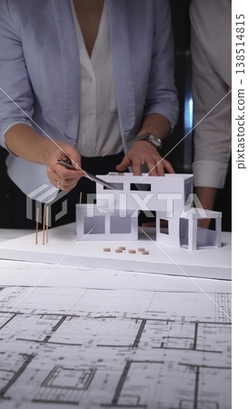 Architects developing a future building project, using architectural plans and a paper model to create a residential structure design. Concept of architecture and business in low key lighting Architects developing a future building project, using architectural plans and a paper model to create a residential structure design. Concept of architecture and business in low key lighting 138514815