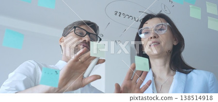 Business colleagues collaborating on strategy, brainstorming ideas for success, writing on a transparent glass board with sticky notes in a modern office Business colleagues collaborating on strategy, brainstorming ideas for success, writing on a transparent glass board with sticky notes in a modern office 138514918