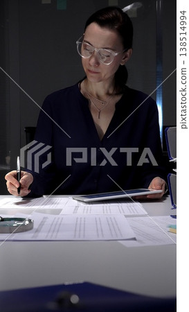 Woman working late, signing documents and analyzing data on a tablet by office lamp light at night. Audit and taxes in business Woman working late, signing documents and analyzing data on a tablet by office lamp light at night. Audit and taxes in business 138514994