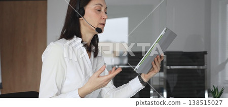 Businesswoman in a white shirt and headset leading a video call from a modern office, presenting with a tablet and gesturing as she discusses strategy and trains remote colleagues Businesswoman in a white shirt and headset leading a video call from a modern office, presenting with a tablet and gesturing as she discusses strategy and trains remote colleagues 138515027