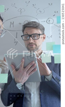 Focused businessman analyzing financial data and brainstorming new project strategies, writing important notes on a transparent glass board wall with sticky notes while holding a tablet Focused businessman analyzing financial data and brainstorming new project strategies, writing important notes on a transparent glass board wall with sticky notes while holding a tablet 138515041