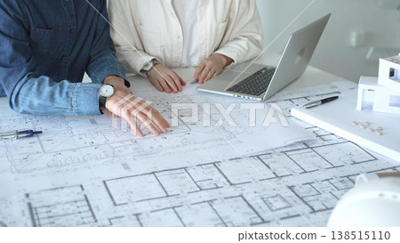 Two architects collaborate over blueprints and a model, using a compass and laptop to measure and plan detailed floor plans, symbolizing teamwork in property development Two architects collaborate over blueprints and a model, using a compass and laptop to measure and plan detailed floor plans, symbolizing teamwork in property development 138515110