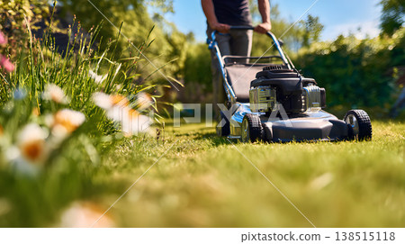 Man operates blue lawn mower trimming green grass in backyard garden. Man mowing lawn in sunny garden backyard landscaping maintenance concept. 138515118