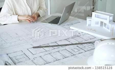 Architect working on new residential project designs, using laptop and reviewing blueprints with an architectural model and safety helmet on planning table 138515128